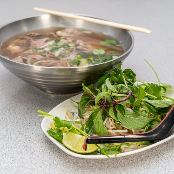 A bowl of Vietnamese pho with beef and noodles, alongside a plate of fresh herbs and lime.