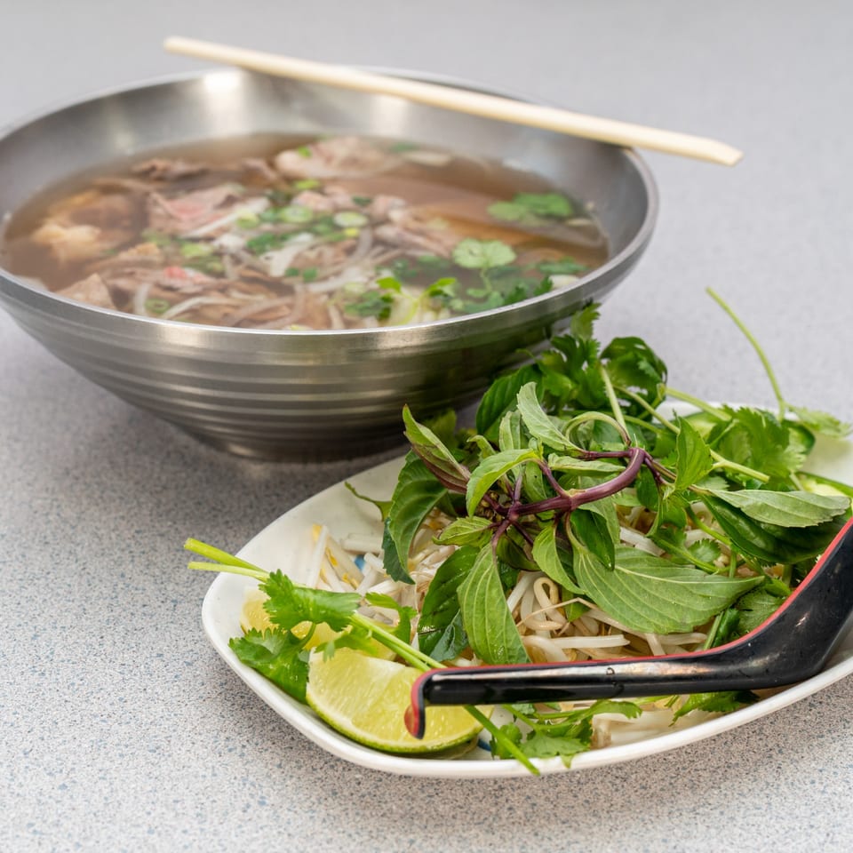A bowl of Vietnamese pho with beef and noodles, alongside a plate of fresh herbs and lime.