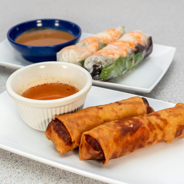 Two plates of Vietnamese spring rolls and egg rolls with dipping sauces on a gray table.