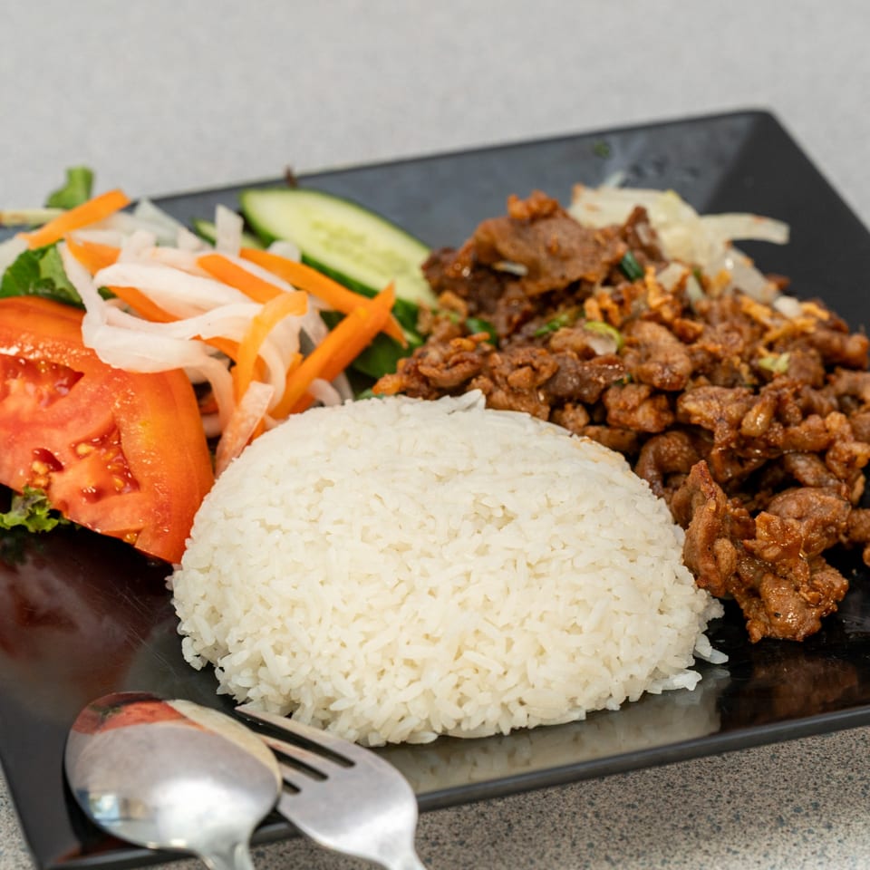 Vietnamese dish with white rice, stir-fried meat, sliced tomatoes, cucumbers, and pickled vegetables.